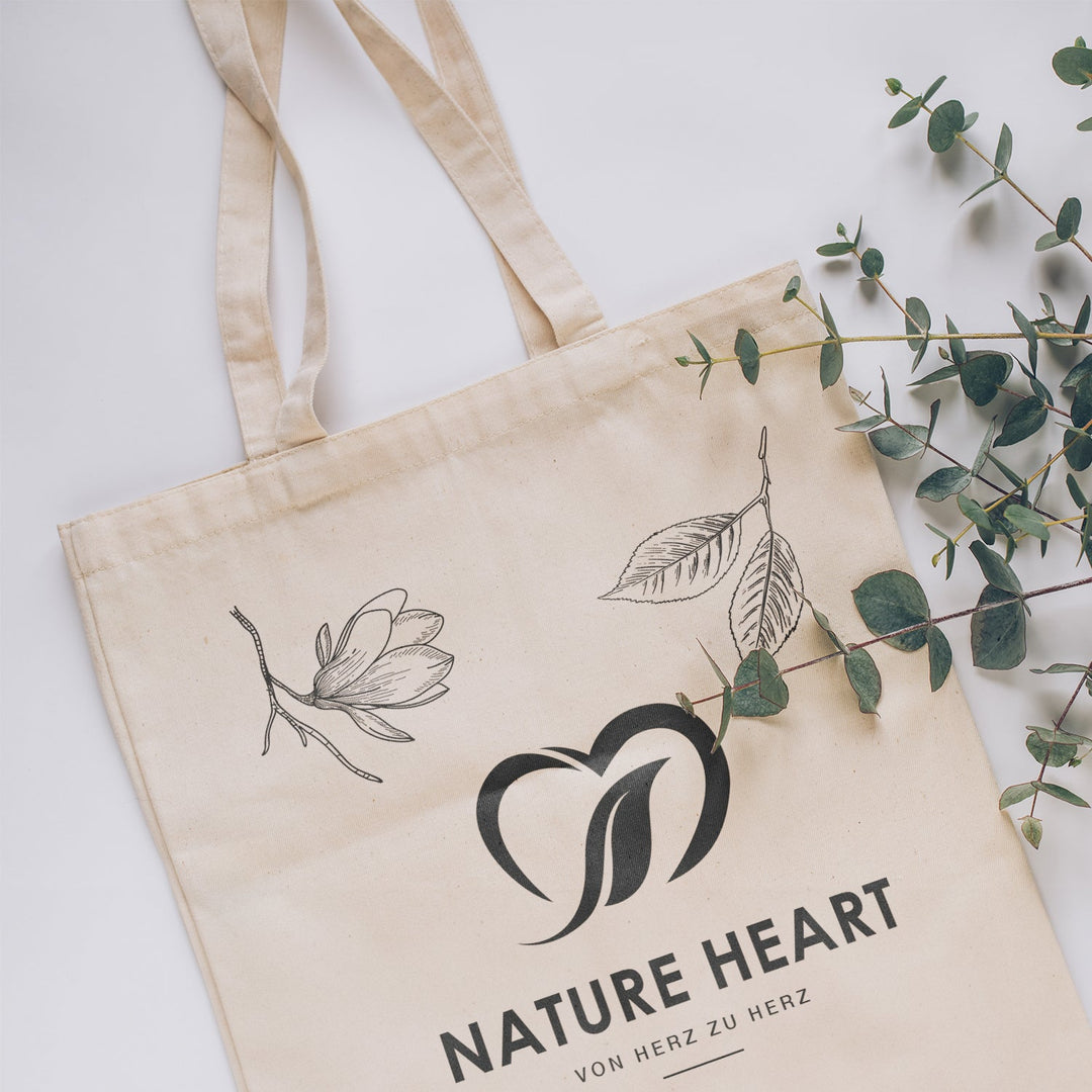 Baumwolltasche Heart flach liegend mit Logo und gezeichnetem Blüten- und Blattmotiv neben grünen Eukalyptuszweigen