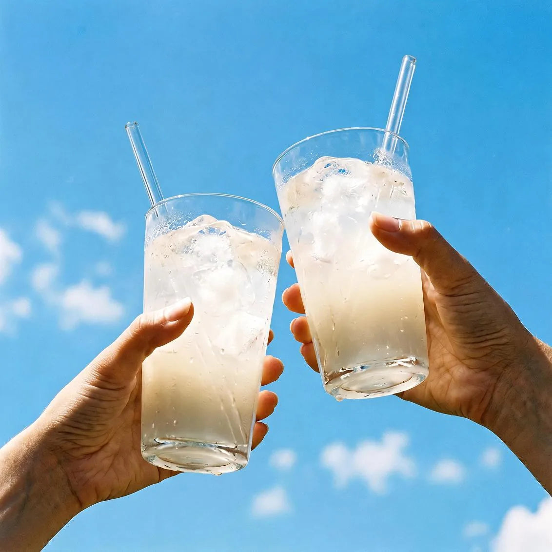 Zwei Hände stoßen vor blauem Himmel mit Gläsern Elektrolyt+ Salty Orange an, mit Eiswürfeln und Glasstrohhalmen