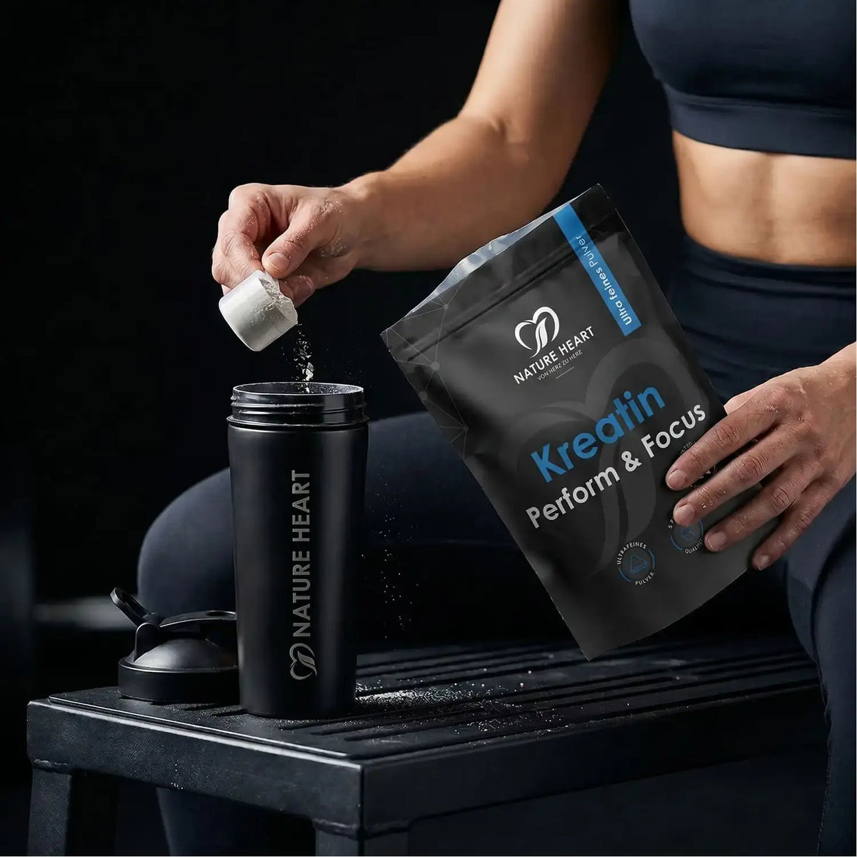 Sportlerin gibt Kreatin Perform & Focus mit dem Messlöffel aus dem Beutel in einen schwarzen Shaker auf einer Trainingsbank