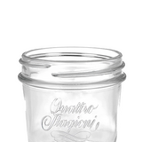 Leeres transparentes Glas mit Schraubrand und eingeprägtem Schriftzug „Quattro Stagioni“, angeschnitten im unteren Bildbereich vor grauem Hintergrund.