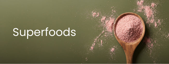 Banner mit der Aufschrift „superfoods“ auf grünem Hintergrund, rechts ein Holzlöffel mit rosa Pulver und verstreutem Pulver.