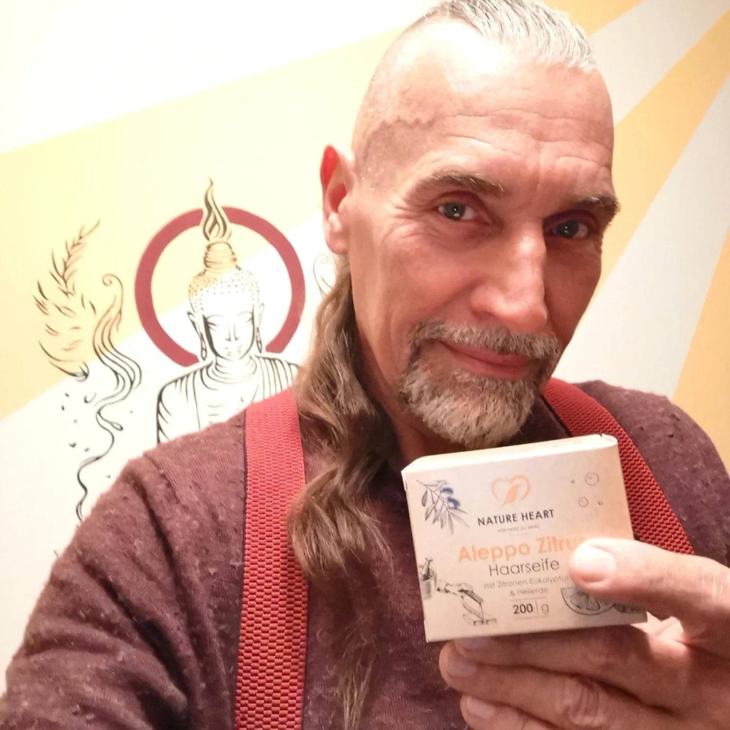 Ein Mann mit grauem Bart präsentiert eine Packung Nature Heart Aleppo Zitrone Haarseife vor einem Hintergrund mit Buddha-Motiv.