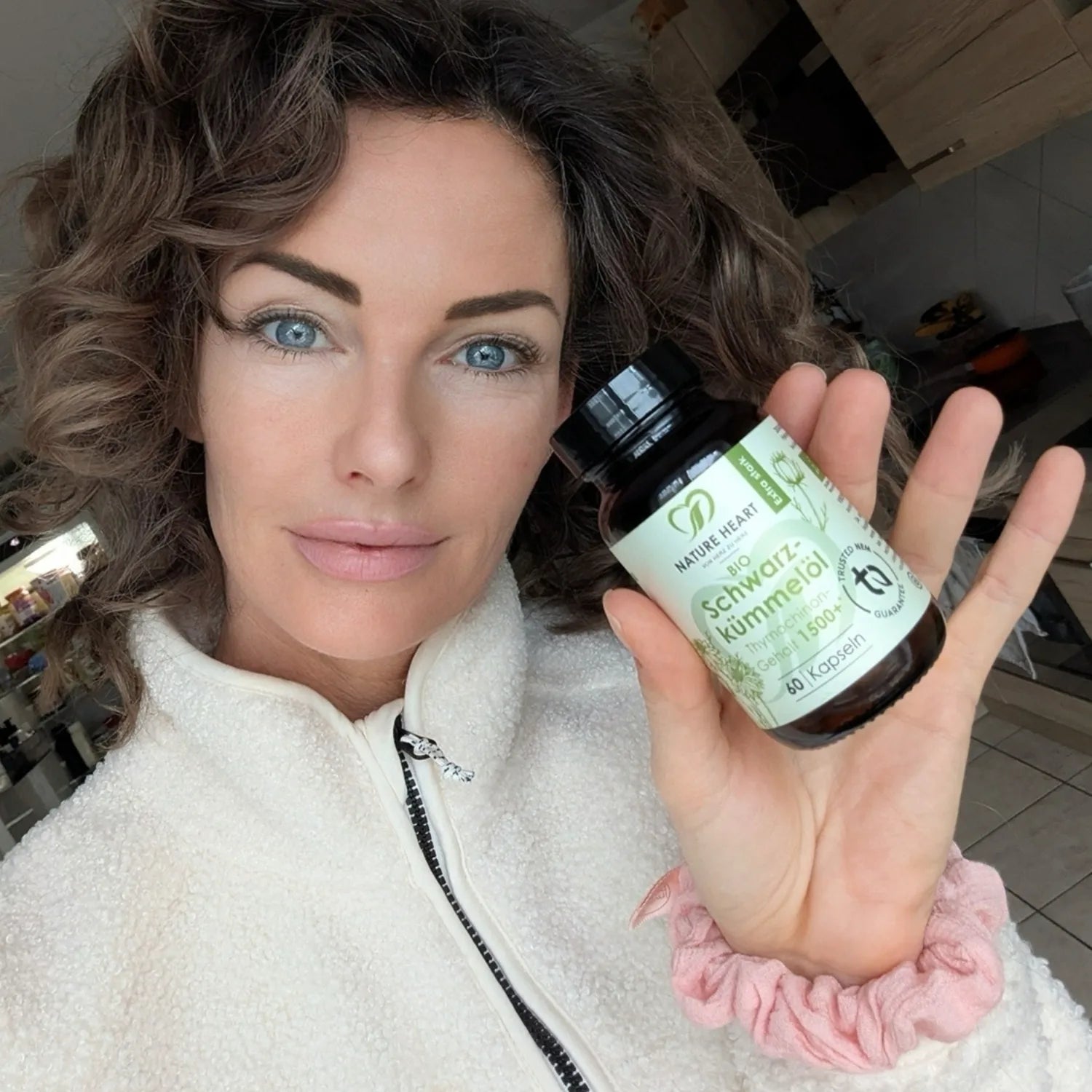 Eine Frau mit lockigen braunen Haaren und blauen Augen zeigt eine Flasche Nature Heart Bio Schwarzkümmelöl Kapseln.