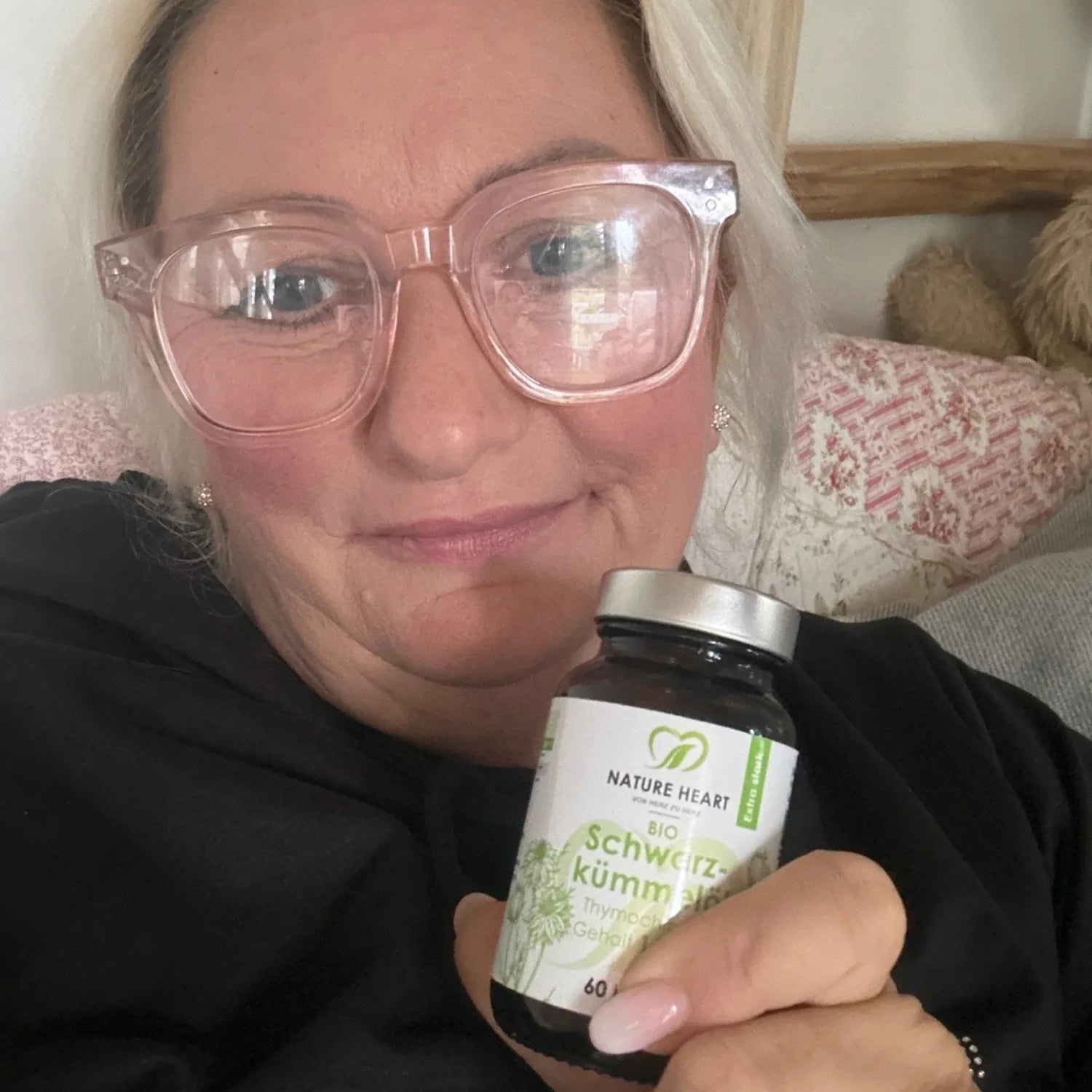 Eine lächelnde Frau mit rosa Brille hält ein Glas Nature Heart Bio Schwarzkümmelöl in die Kamera.