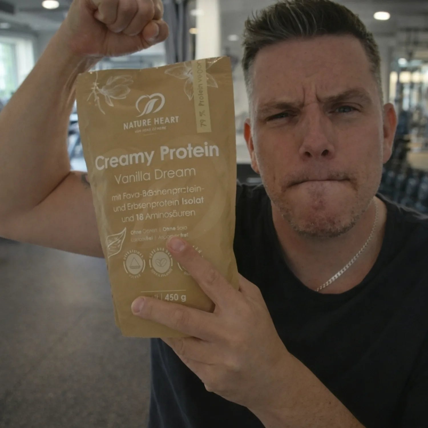 Ein Mann im Fitnessstudio spannt seinen Bizeps an und hält einen Beutel Nature Heart Creamy Protein Vanilla Dream Pulver.