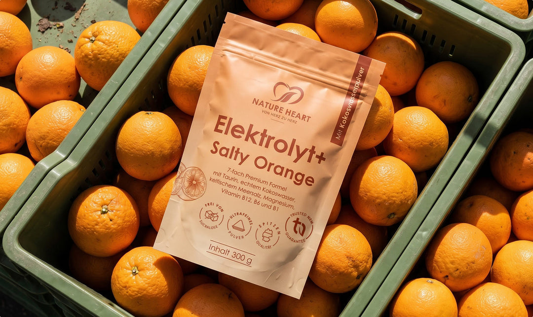 Eine Packung Nature Heart Elektrolyt+ Salty Orange liegt inmitten einer grünen Kiste voller frischer, reifer Orangen im hellen Tageslicht.