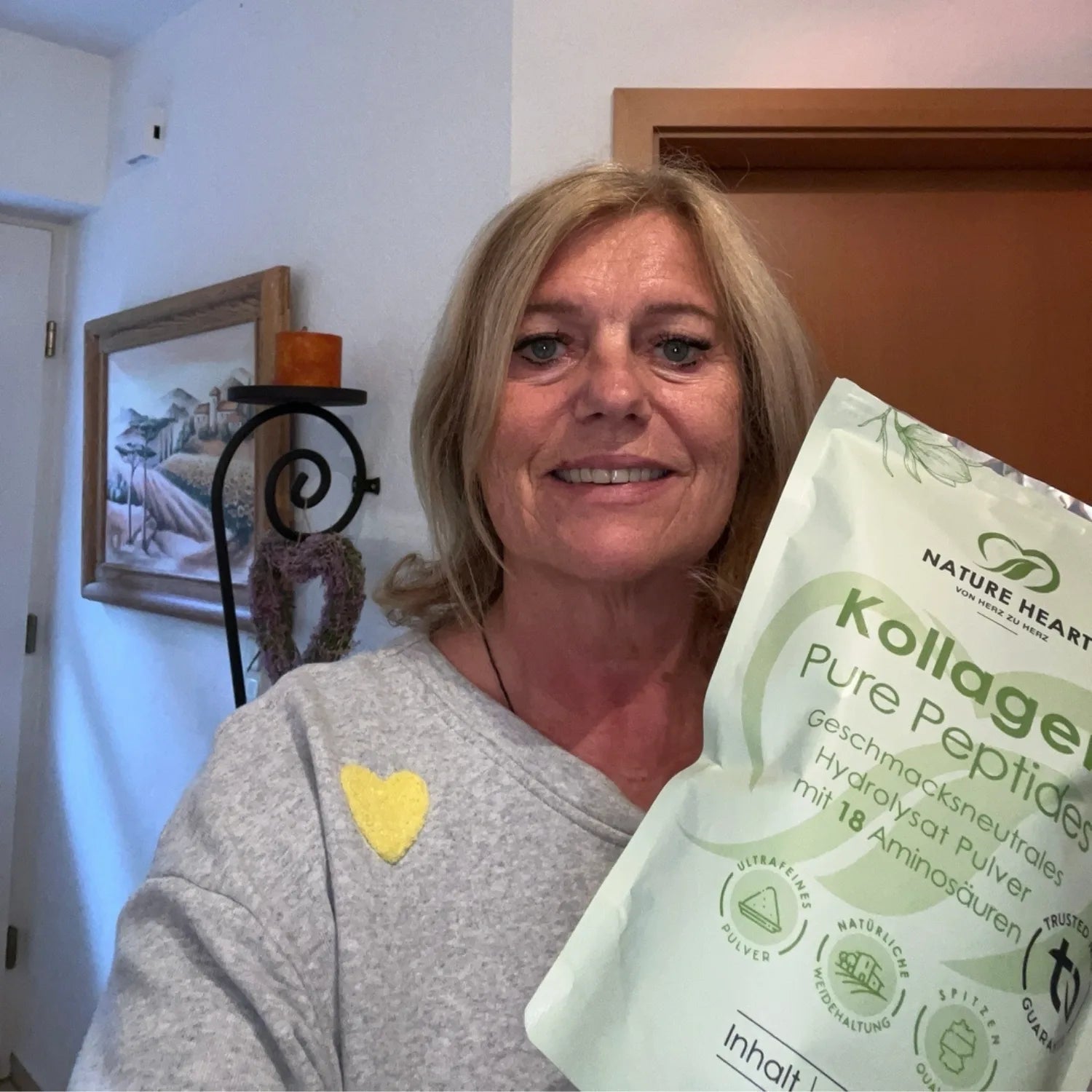 Eine lächelnde Frau im grauen Pullover hält einen großen Beutel Nature Heart Kollagen Pure Peptides Pulver.