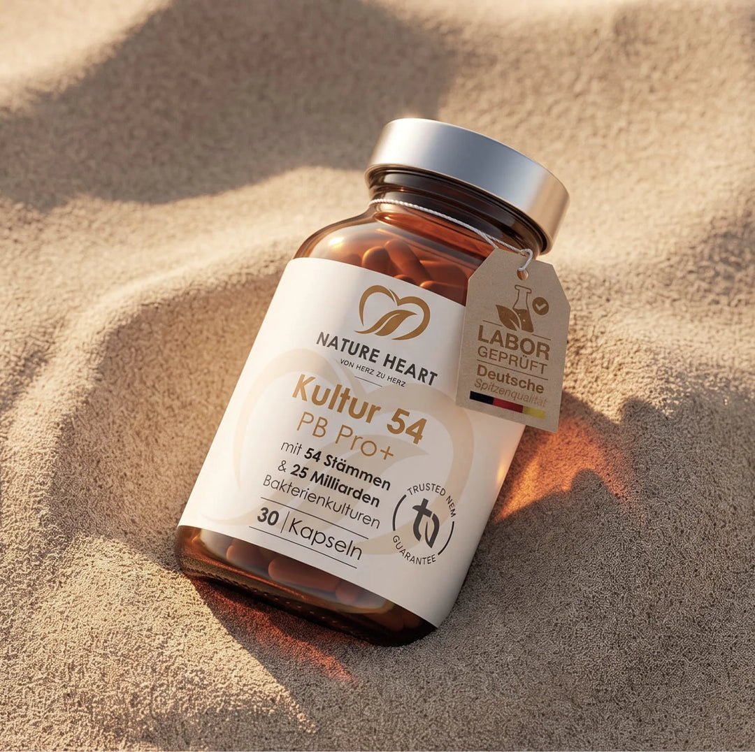 Eine Braunglasflasche Kultur 54 PB Pro+ Kapseln von Nature Heart liegt im sanften Dünensand. Das Etikett verweist auf 54 Stämme und 25 Milliarden Bakterienkulturen für die Reiseapotheke.