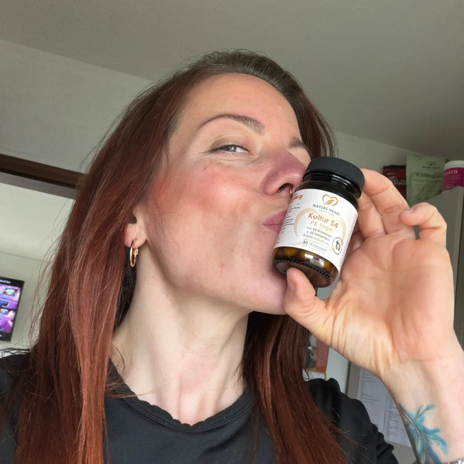 Eine rothaarige Frau spitzt die Lippen für einen Kuss in Richtung eines kleinen Glases Nature Heart Kultur 54 PB Pro+ Kapseln.