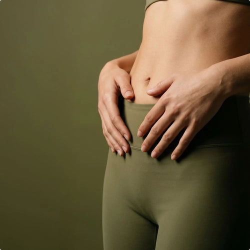 Eine Frau in sportlicher, grüner Kleidung hält schützend und sanft beide Hände auf ihren entspannten Bauch.