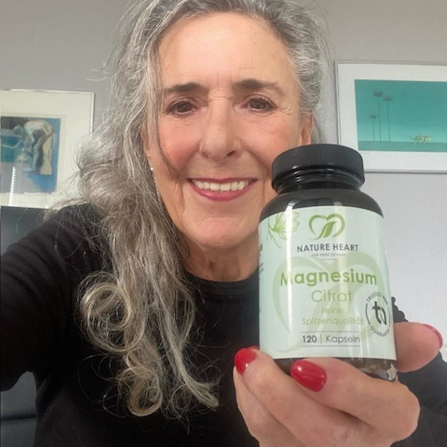 Eine lächelnde Frau mit langen grauen Haaren hält ein Glas Nature Heart Magnesium Citrat Kapseln.