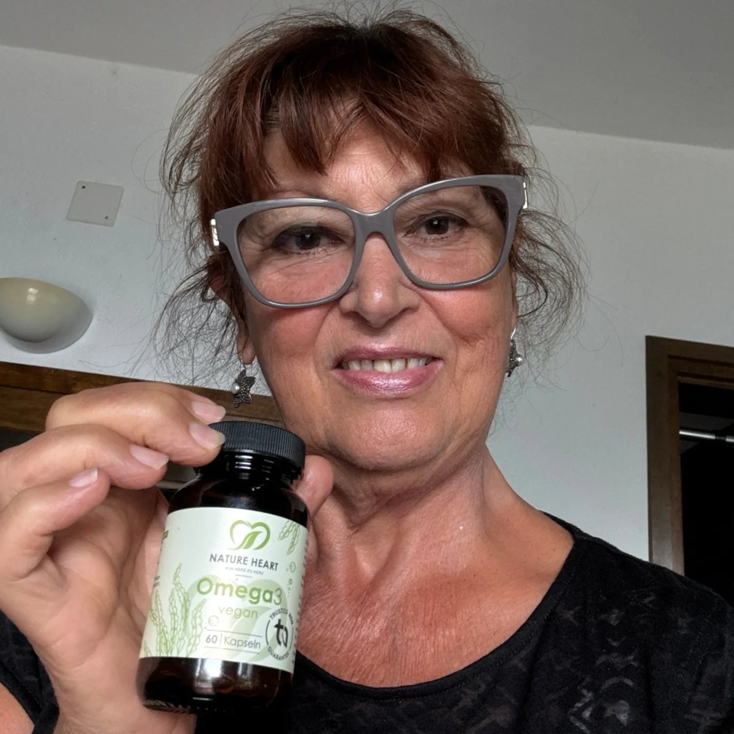 Eine lächelnde Frau mit Brille präsentiert eine Dose Nature Heart Omega 3 Vegan Kapseln.