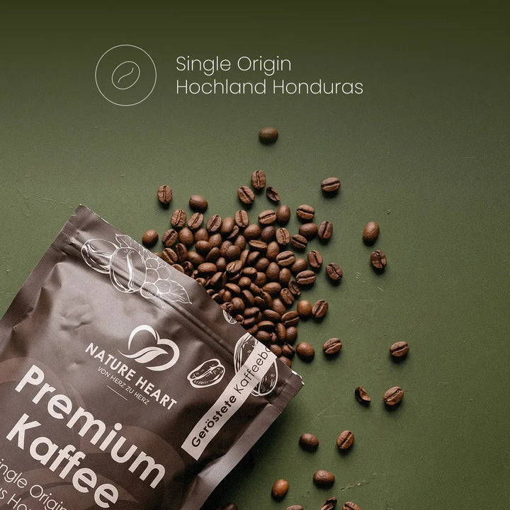 Eine Kaffeeverpackung von Nature Heart Premium Kaffee liegt auf grünem Untergrund neben verstreuten, gerösteten Bohnen unter dem Schriftzug „Single Origin Hochland Honduras“.