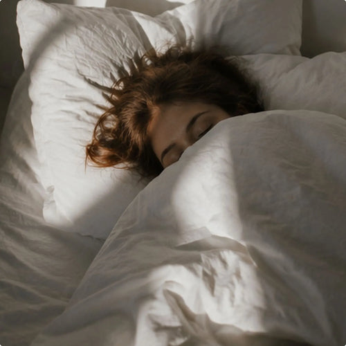 Eine Person liegt gemütlich eingekuschelt unter einer weißen Bettdecke im Bett, während morgendliche Sonnenstrahlen durch das Fenster auf das Kissen fallen.