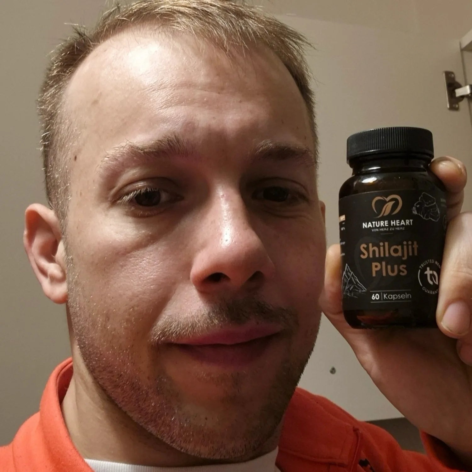 Ein Mann blickt in die Kamera und hält ein Glas Nature Heart Shilajit Plus Kapseln neben sein Gesicht.