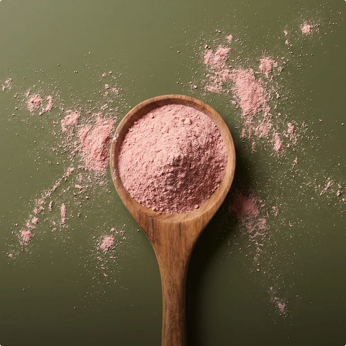Ein rustikaler Holzlöffel, der reichlich mit fein gemahlenem, rosafarbenem Superfood-Pulver befüllt ist, fotografiert aus der Vogelperspektive auf einem olivgrünen Hintergrund mit dekorativ verstreuten Pulverresten.