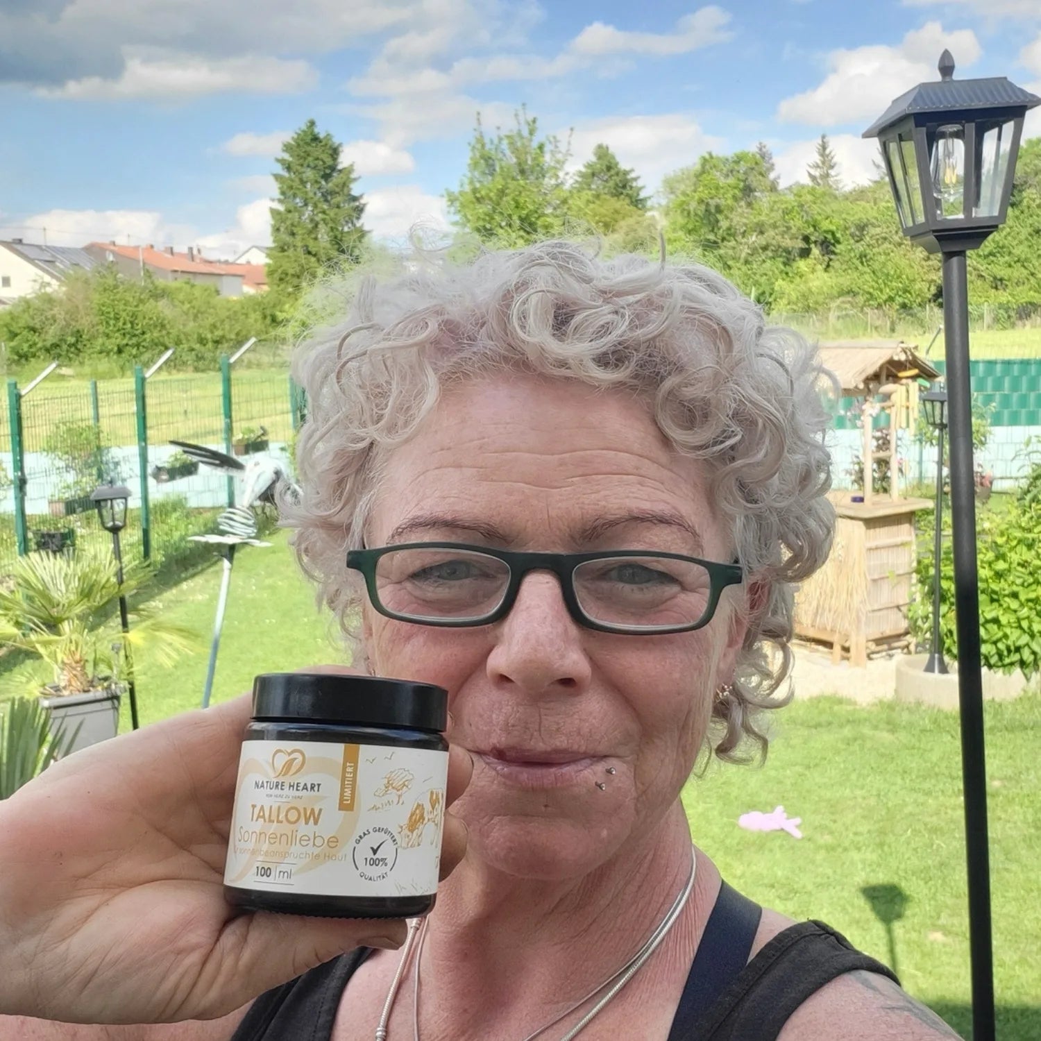 Eine Frau mit kurzen lockigen Haaren und Brille hält im Garten einen Tiegel Nature Heart Tallow Sonnenliebe in die Kamera.
