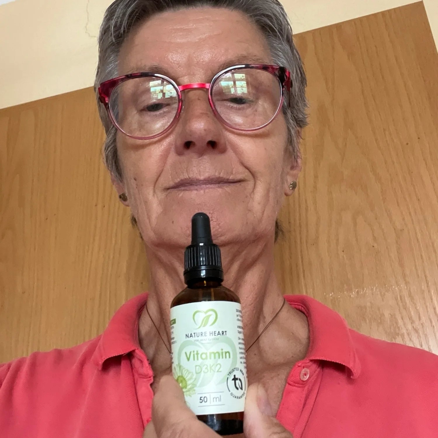 Eine Frau mit Brille hält eine Pipettenflasche Nature Heart Vitamin D3K2 Tropfen vor sich.