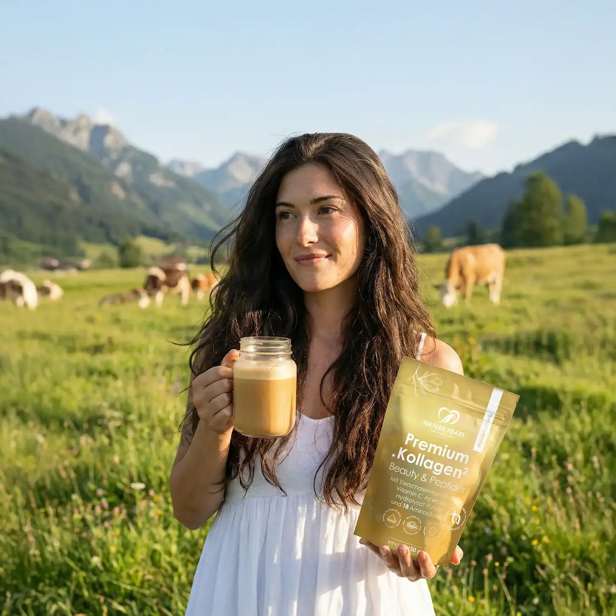 Frau mit Kollagen-Drink im Glas und einem Beutel Premium Kollagen auf einer Bergwiese mit grasenden Rindern
