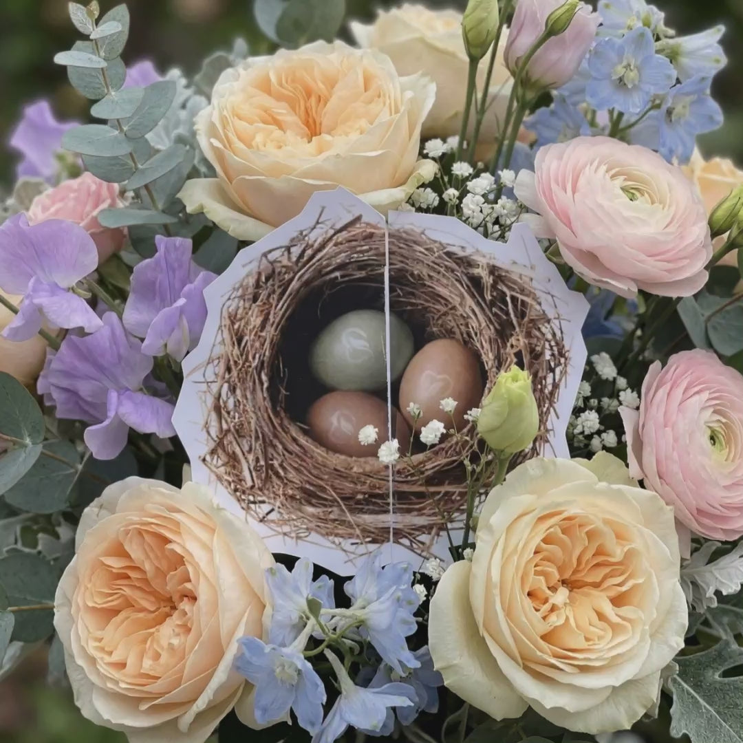 Das Video zeigt ein osterliches Blumenarrangement mit pastellfarbenen Blüten und einem Nest mit Eiern, das anschließend als Motiv auf einer geöffneten Nature Heart Geschenkkarte in grünen Pflanzen zu sehen ist.