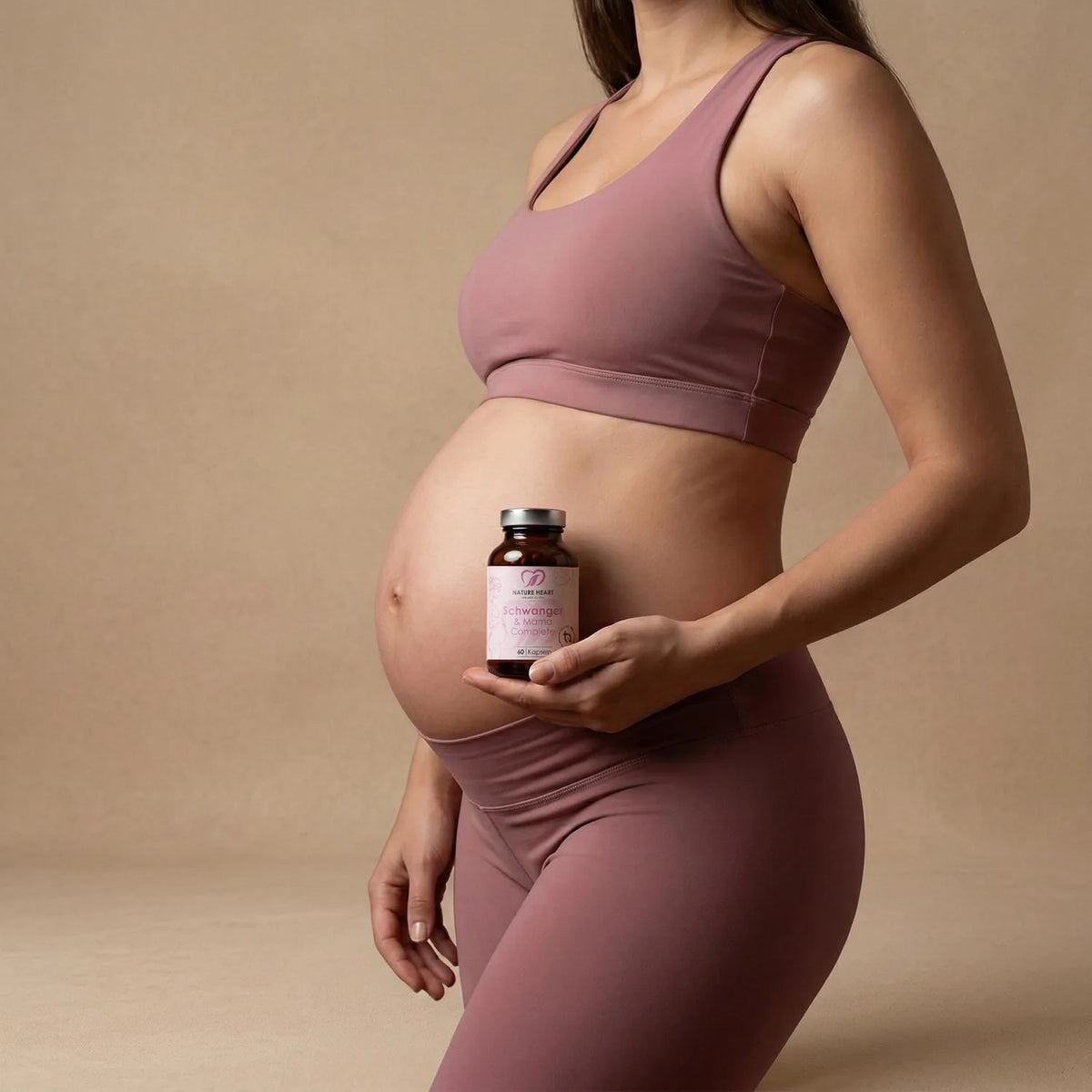 Schwangere Frau in altrosa Sportkleidung hält ein Glas Schwanger & Mama Complete mit 60 Kapseln vor ihren Babybauch
