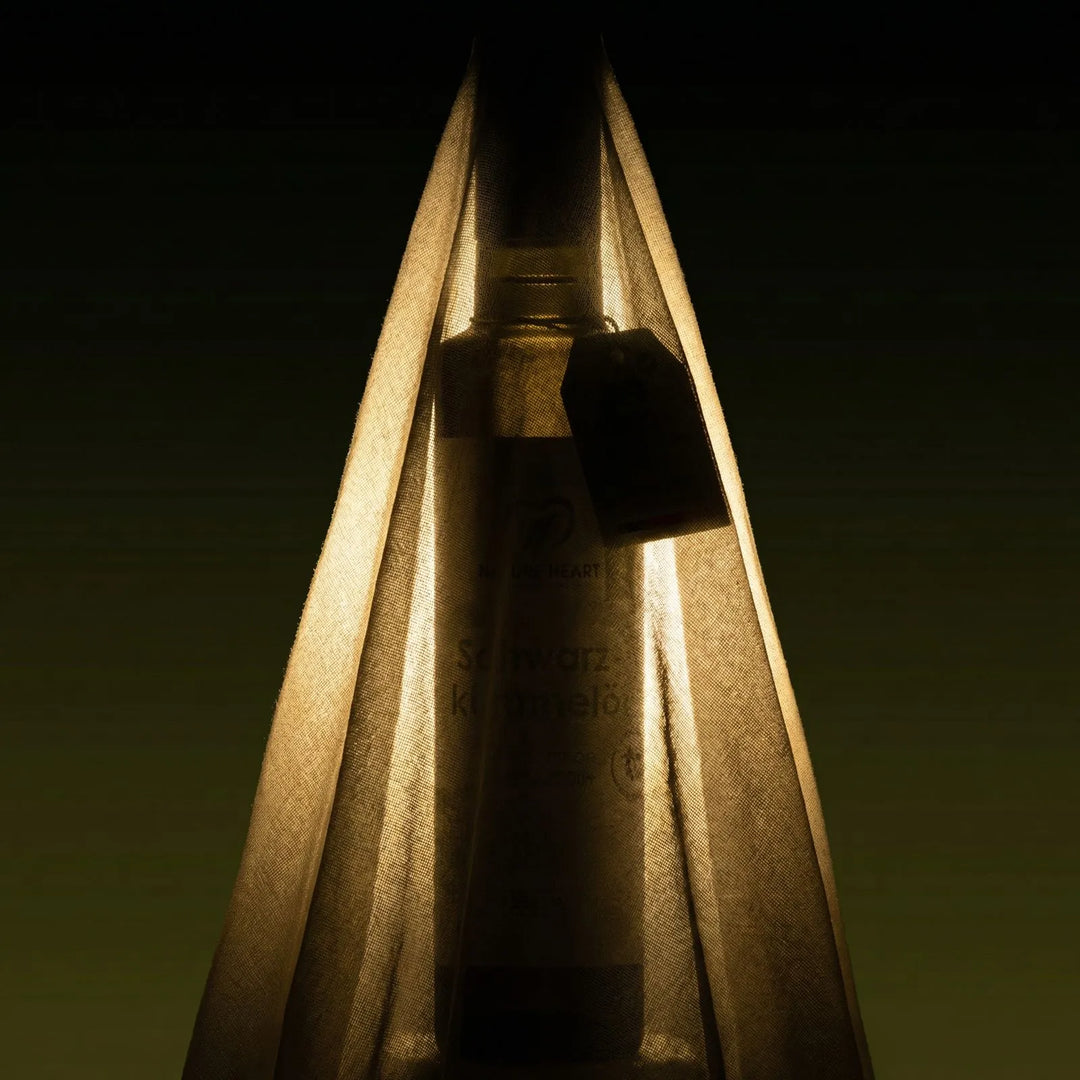 Dunkel inszenierte Glasflasche mit Nature Heart Schwarzkümmelöl hinter halbtransparentem Stoff, mit warmem Licht und sichtbarem Tag am Flaschenhals.